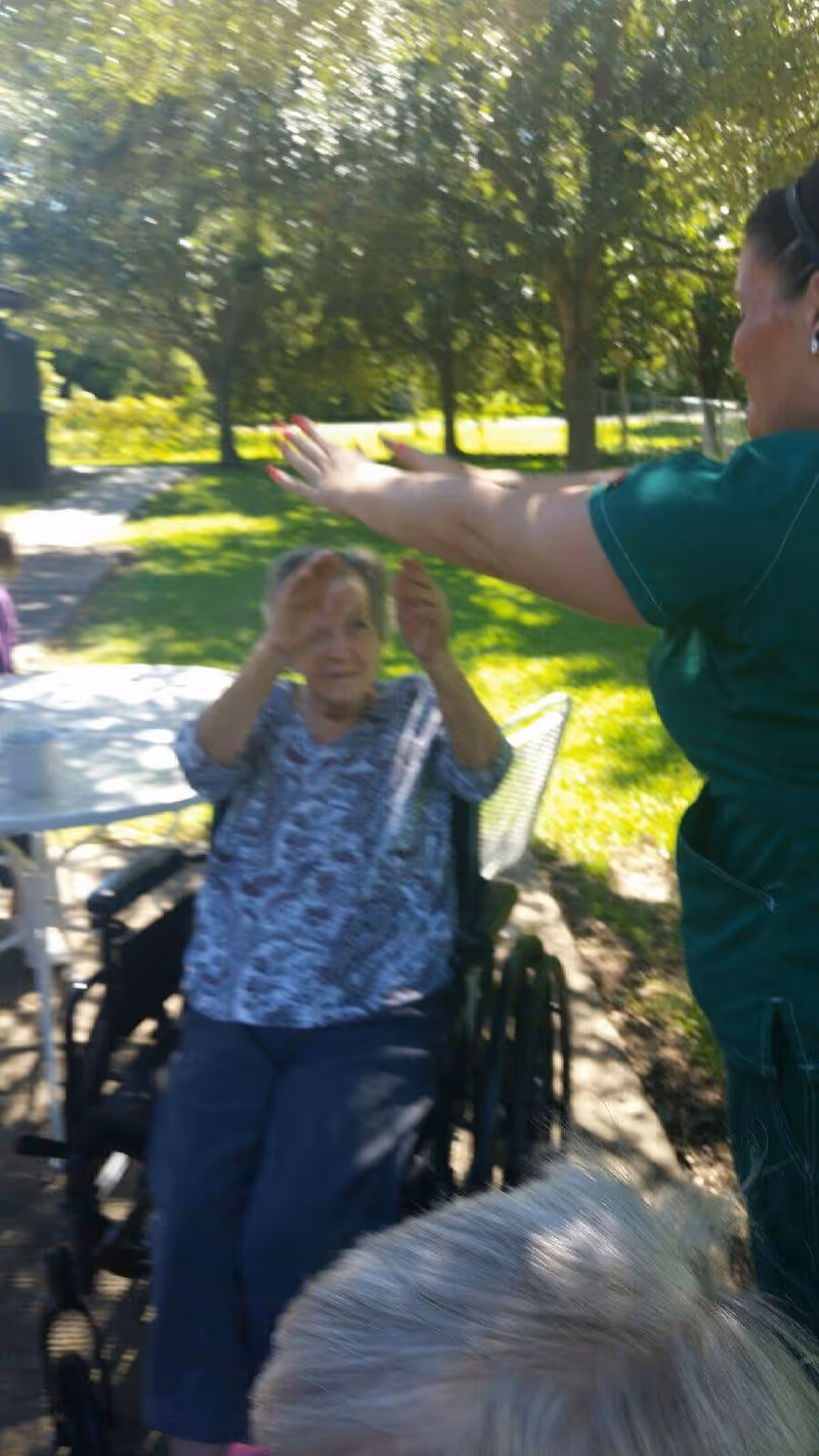 An elderly woman sitting in a wheelchair outdoors, raising her hands towards a caregiver in green scrubs who is reaching out to her. They are in a sunny garden area with trees and grass in the background.