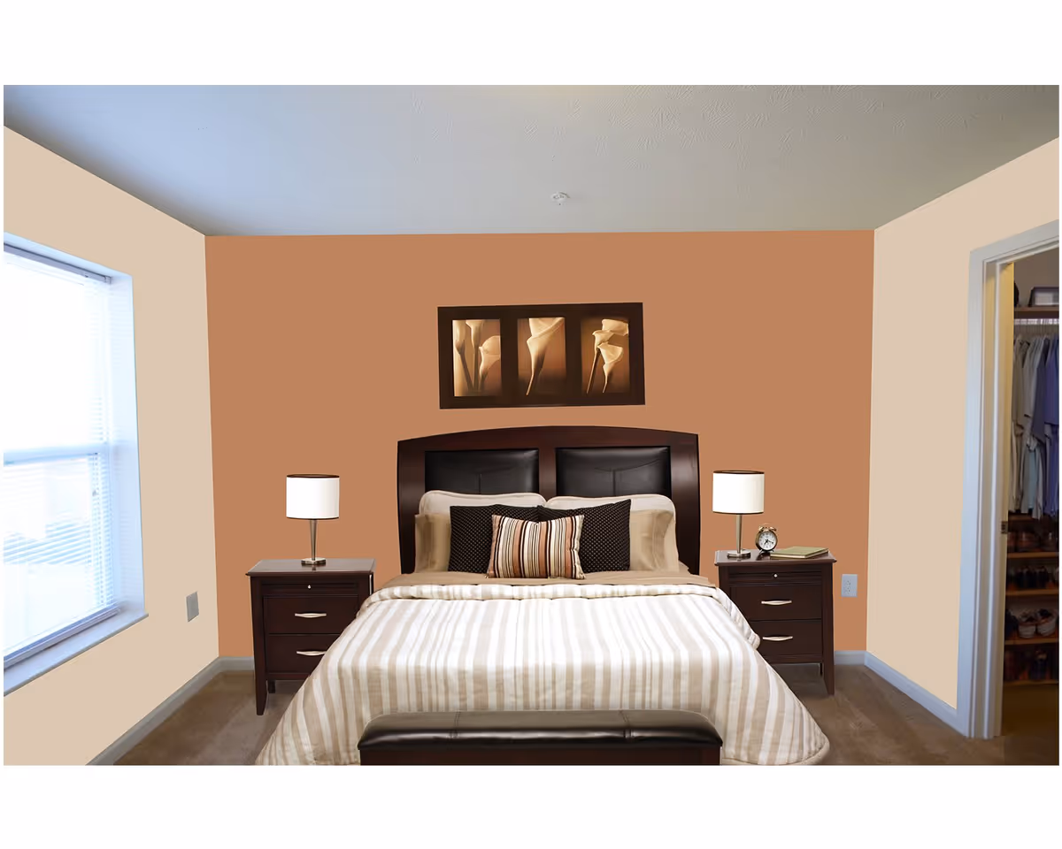 A bedroom with a bed centered against a two-tone painted wall. The bed has a dark wooden headboard with black cushioned panels, beige and brown striped bedding, and multiple pillows. On each side of the bed are dark wooden nightstands with white lamps. Above the bed hangs a triptych artwork featuring abstract floral designs. To the right, there is an open doorway leading to a closet with hanging clothes and shoes. A window with blinds is on the left wall.