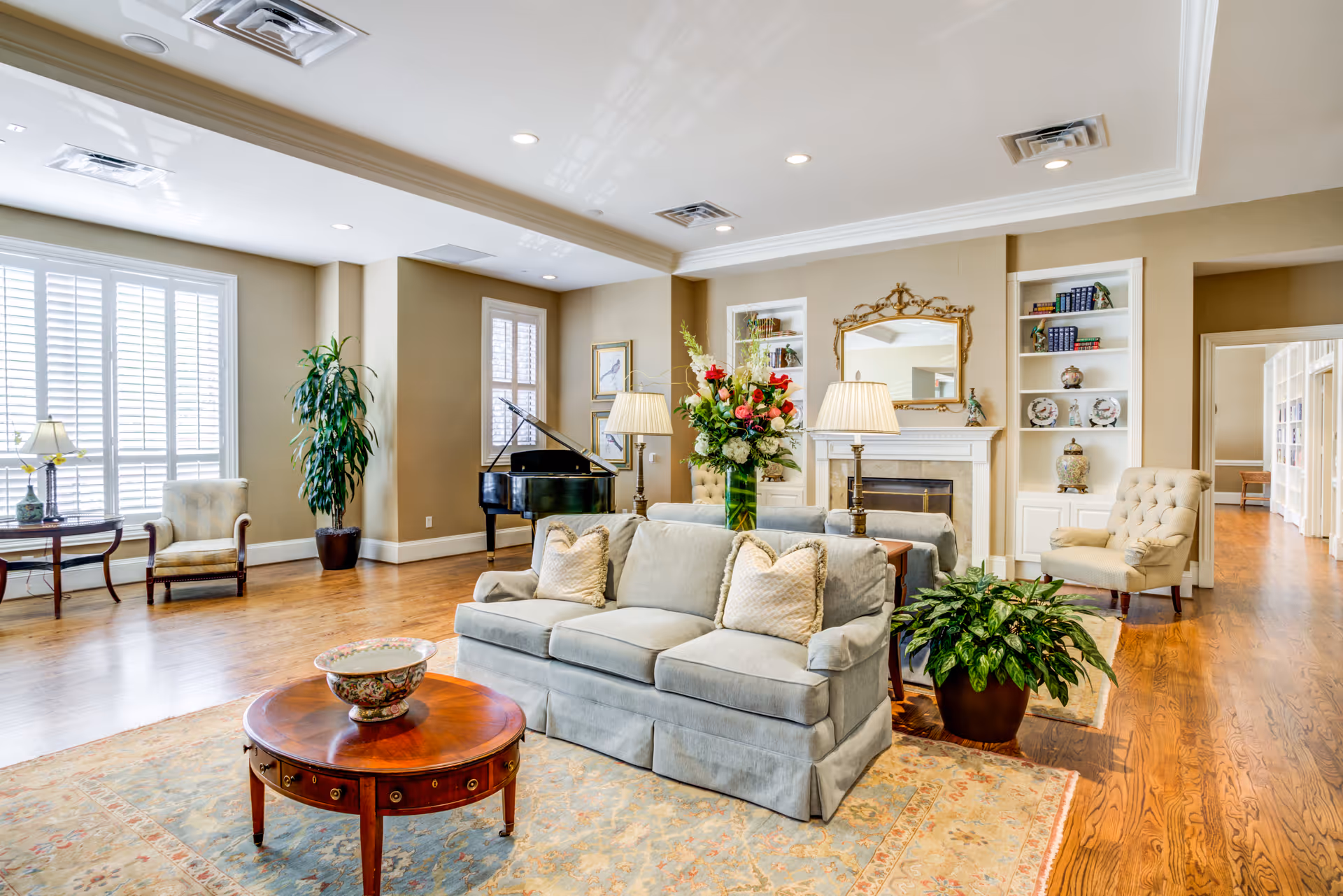 A bright and spacious living room with wooden floors, a light blue sofa with pillows, a round wooden coffee table with a decorative bowl, two armchairs, a grand piano, a fireplace with a gold-framed mirror above it, built-in shelves with decorative items, two table lamps, and several plants. Large windows with white shutters allow natural light to fill the room.