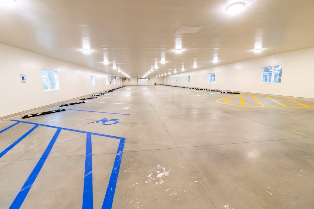 Indoor parking garage with marked parking spaces including a handicapped spot with blue lines and a yellow-striped no parking zone. The garage has white walls, several windows, and ceiling lights evenly spaced along the length of the space.