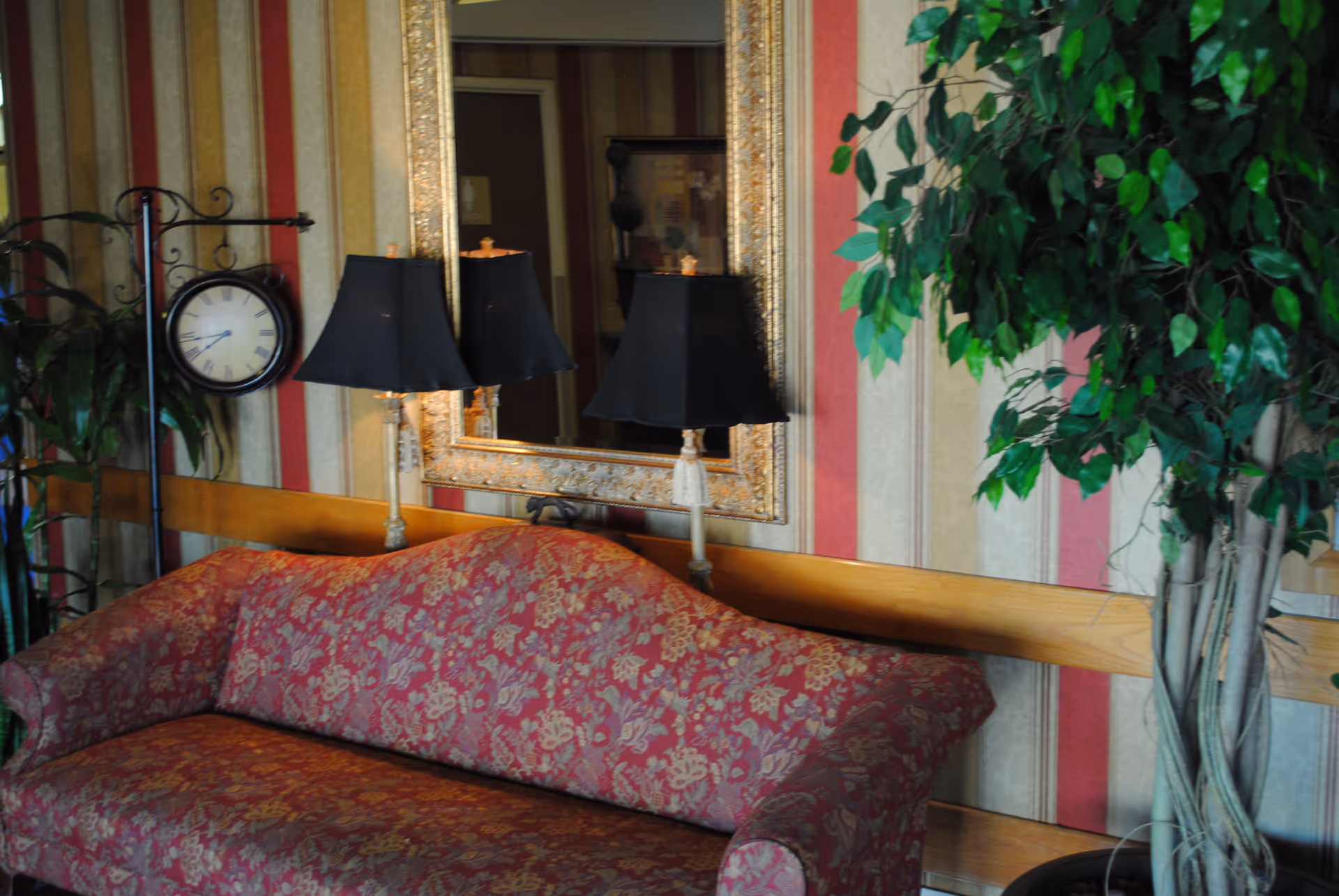 A cozy living room area with a red floral patterned sofa, two black lamps with gold bases, a large ornate gold-framed mirror on the wall, a round clock on a stand, and a tall green leafy plant. The wall behind has vertical striped wallpaper in shades of red, beige, and gold.
