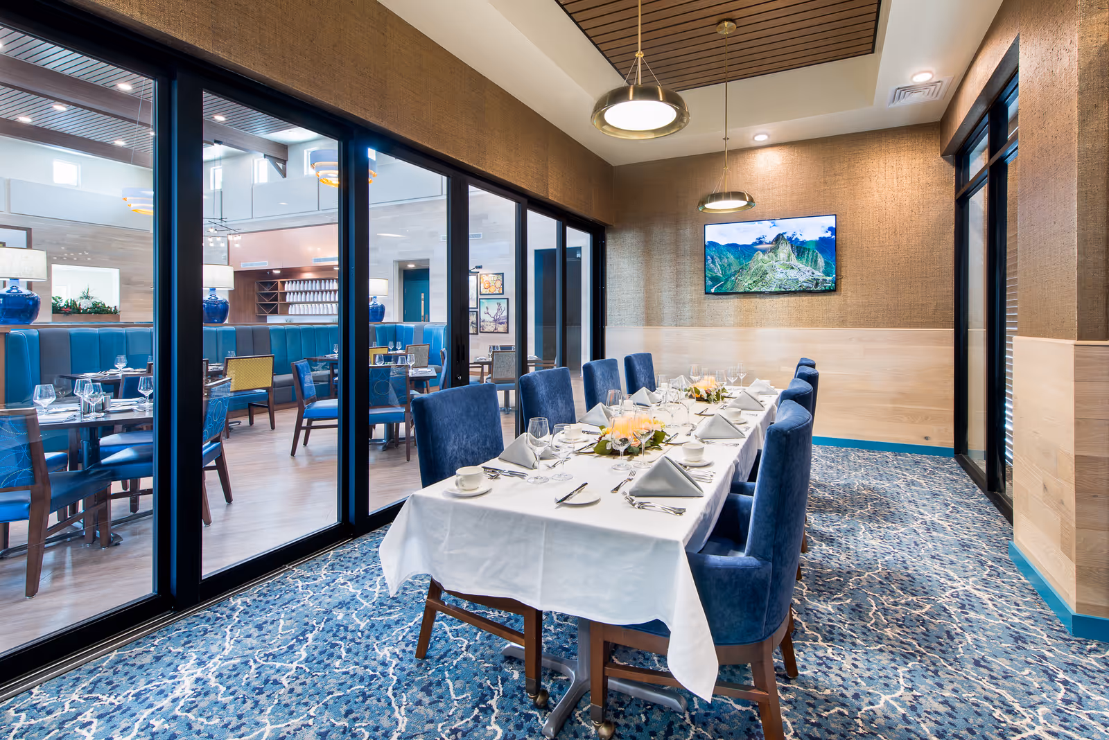 A private dining room with a long table set for a meal, featuring blue upholstered chairs, white tablecloth, folded napkins, glassware, and floral centerpieces. The room has a patterned blue carpet, beige walls with wood paneling, and modern pendant lights. Large glass sliding doors separate this room from a larger dining area with additional tables and chairs.