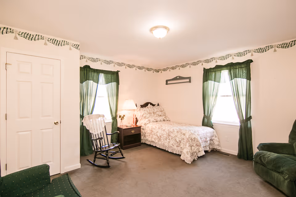 A cozy bedroom with a single bed covered in floral bedding, two windows with green curtains, a wooden rocking chair with a white lace cover, a nightstand with a lamp and flowers, and a green upholstered chair. The walls are white with a decorative green and white border near the ceiling.