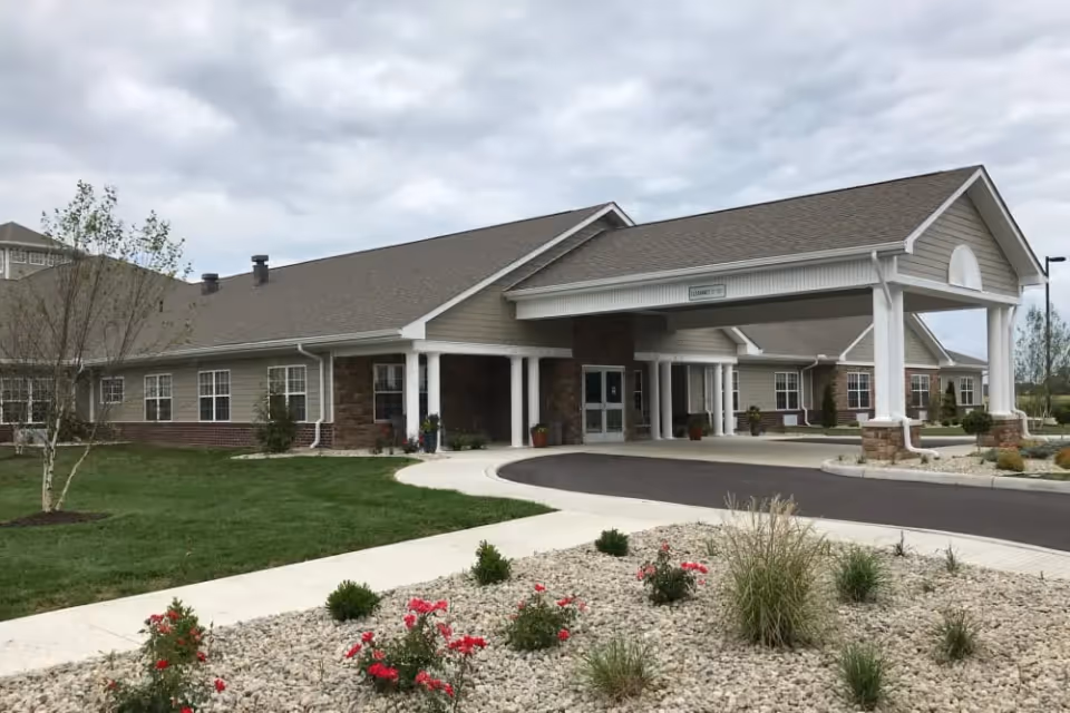 Front view of a single-story senior living building with a covered porte-cochère entrance, driveway, and landscaped grounds.
