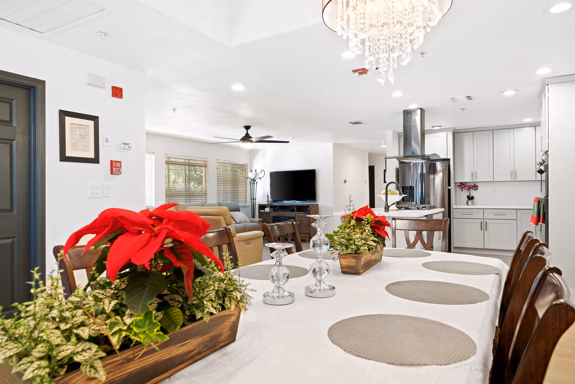 A bright and spacious open-concept dining and living area in a senior living facility. The foreground features a dining table covered with a white tablecloth, decorated with two wooden planters containing red poinsettias and green foliage, and two glass candle holders. The background shows a cozy living room with beige sofas, a flat-screen TV, and large windows with blinds. To the right is a modern kitchen with white cabinetry, stainless steel appliances, and a kitchen island with a sink and faucet. A chandelier hangs above the dining table.