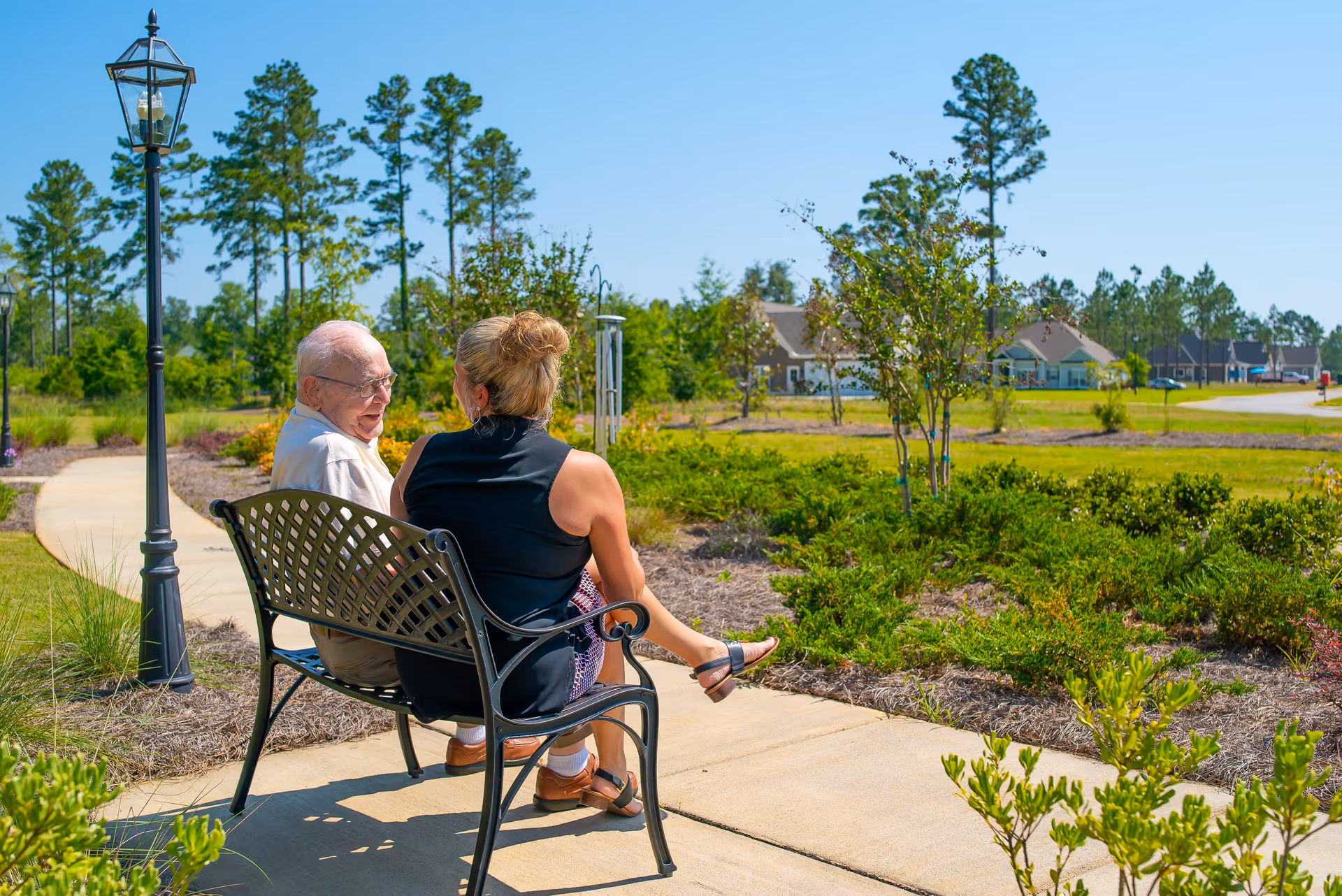 An elderly man and a woman sitting on a black metal bench along a paved pathway in a landscaped outdoor area with green shrubs, trees, and houses in the background under a clear blue sky.
