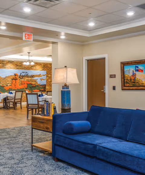 Interior view of a senior living facility showing a blue couch with a cylindrical pillow, a wooden side table with a blue lamp and books, and a dining area in the background with tables and chairs. The walls are beige with a framed painting and a mural on a brick wall that reads 'Light the Lighthouse'. Ceiling lights and an exit sign are visible.