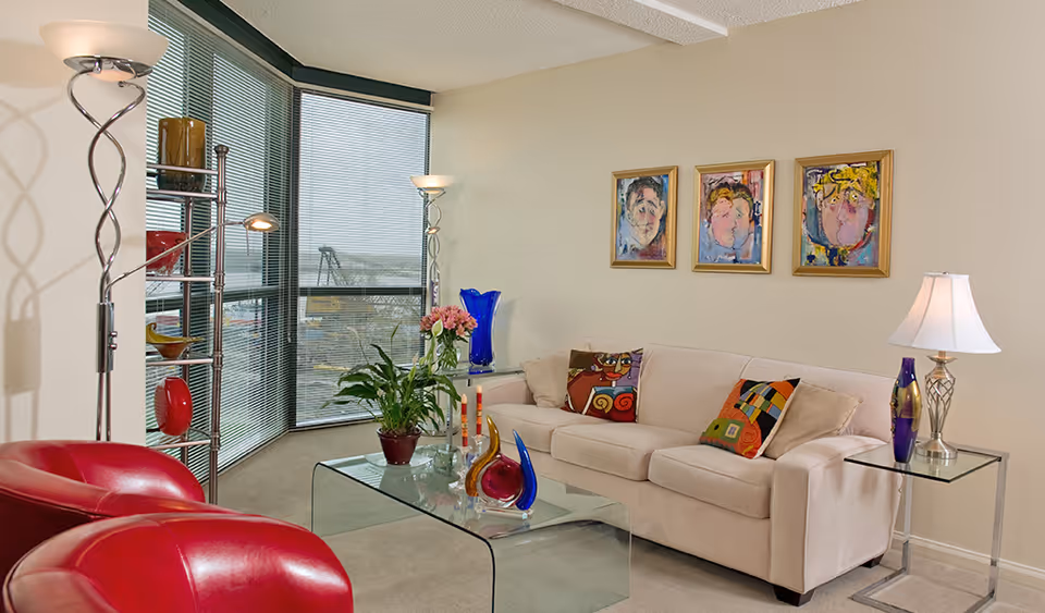 Contemporary living room with a beige sofa, glass coffee table, red chairs, colorful artwork and large corner windows.