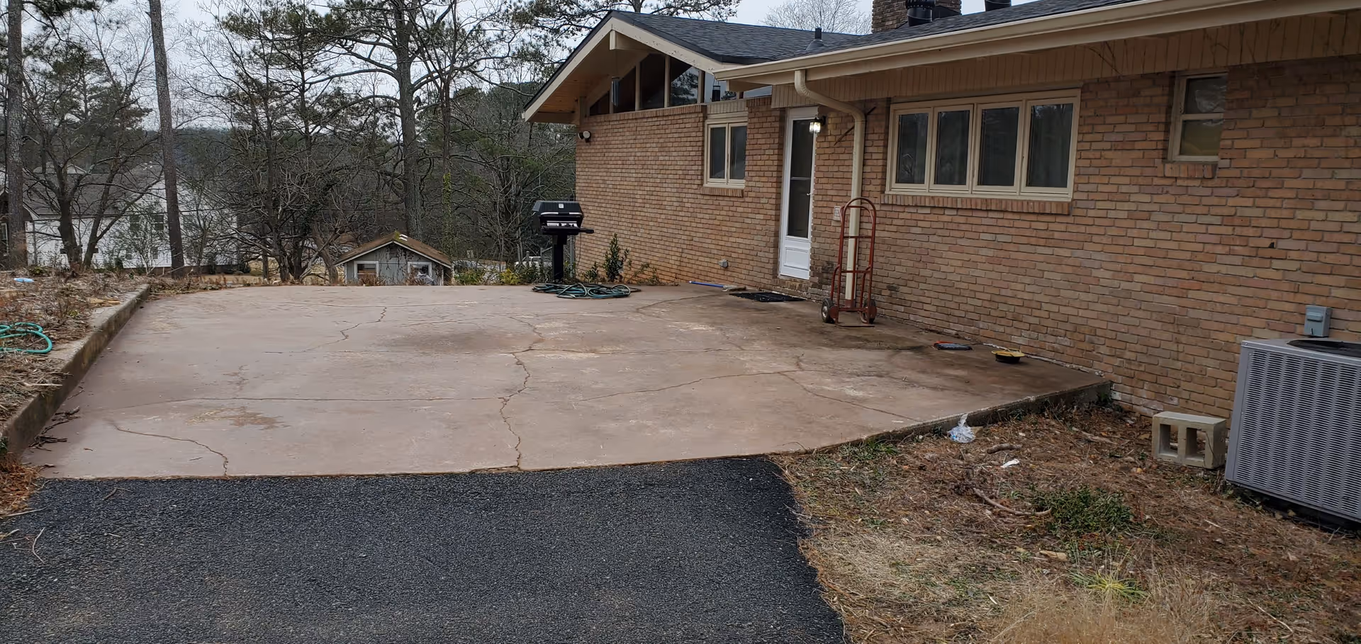 Outdoor concrete patio area adjacent to a brick building with a white door and several windows. There is a black grill, a red hand truck, and an air conditioning unit visible. Surrounding the patio are trees and some dry grass and dirt patches.