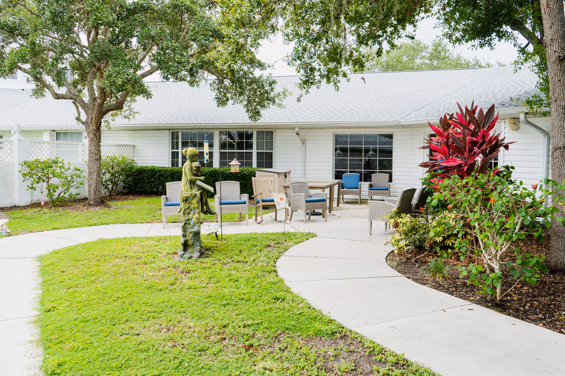 Outdoor patio area at Heron House featuring a curved concrete walkway, green grass, a bronze statue of a girl reading a book, several wicker chairs with blue cushions, a table, and lush plants including a large tree and vibrant red foliage near a white building with large windows.