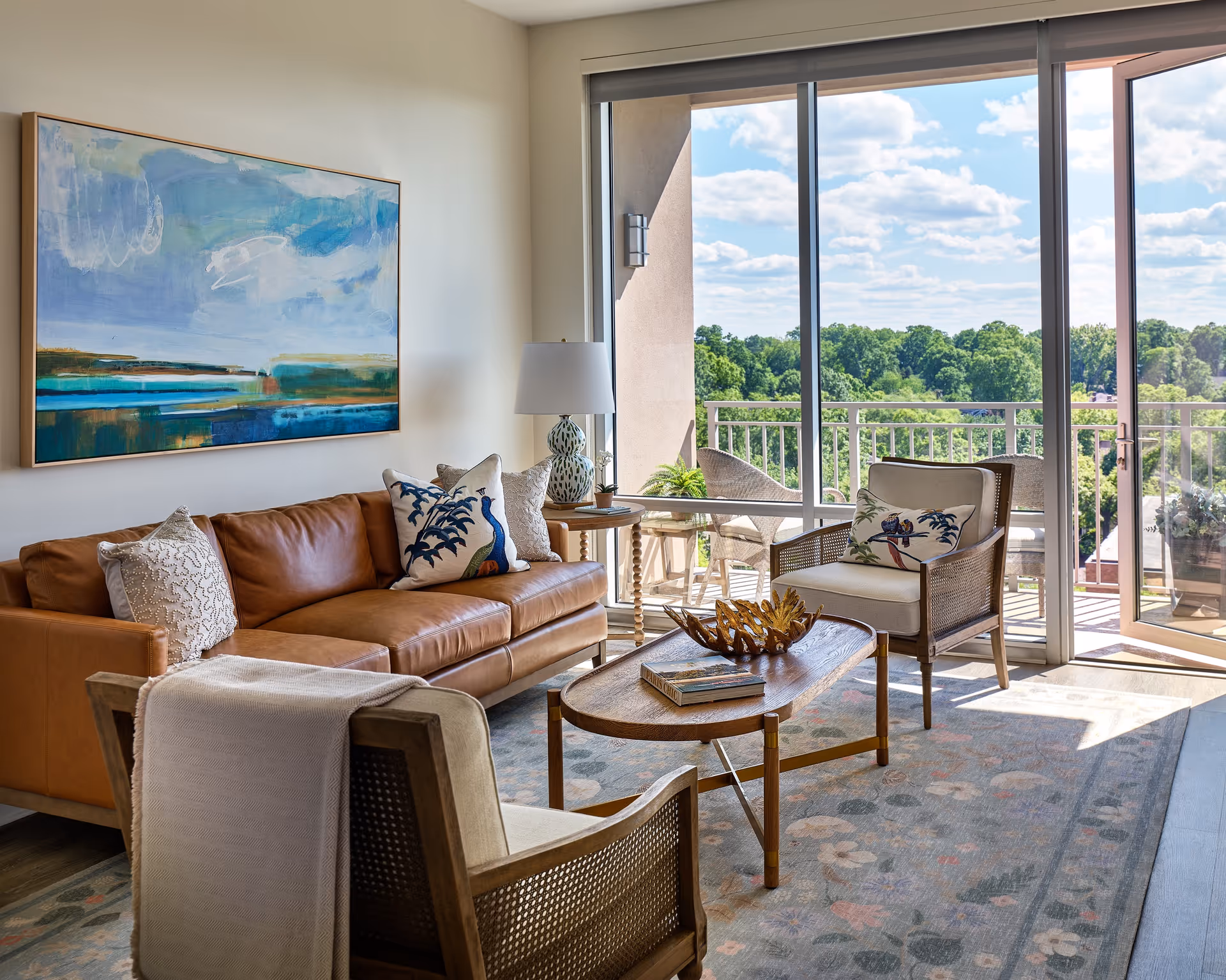 A bright living room with a brown leather sofa adorned with decorative pillows, two cushioned armchairs, a wooden coffee table with books and a decorative piece, a side table with a lamp, and a large abstract painting on the wall. Large sliding glass doors open to a balcony with outdoor seating and a view of green trees under a partly cloudy sky.