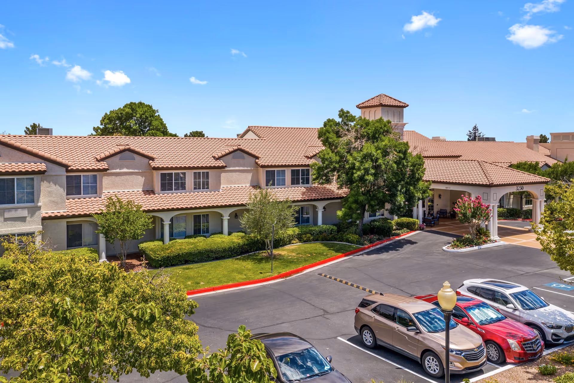 Exterior view of Oakmont of the Lakes senior living facility showing a two-story building with a tiled roof, surrounded by trees and shrubs. There is a parking lot in front with several parked cars and a covered entrance area.