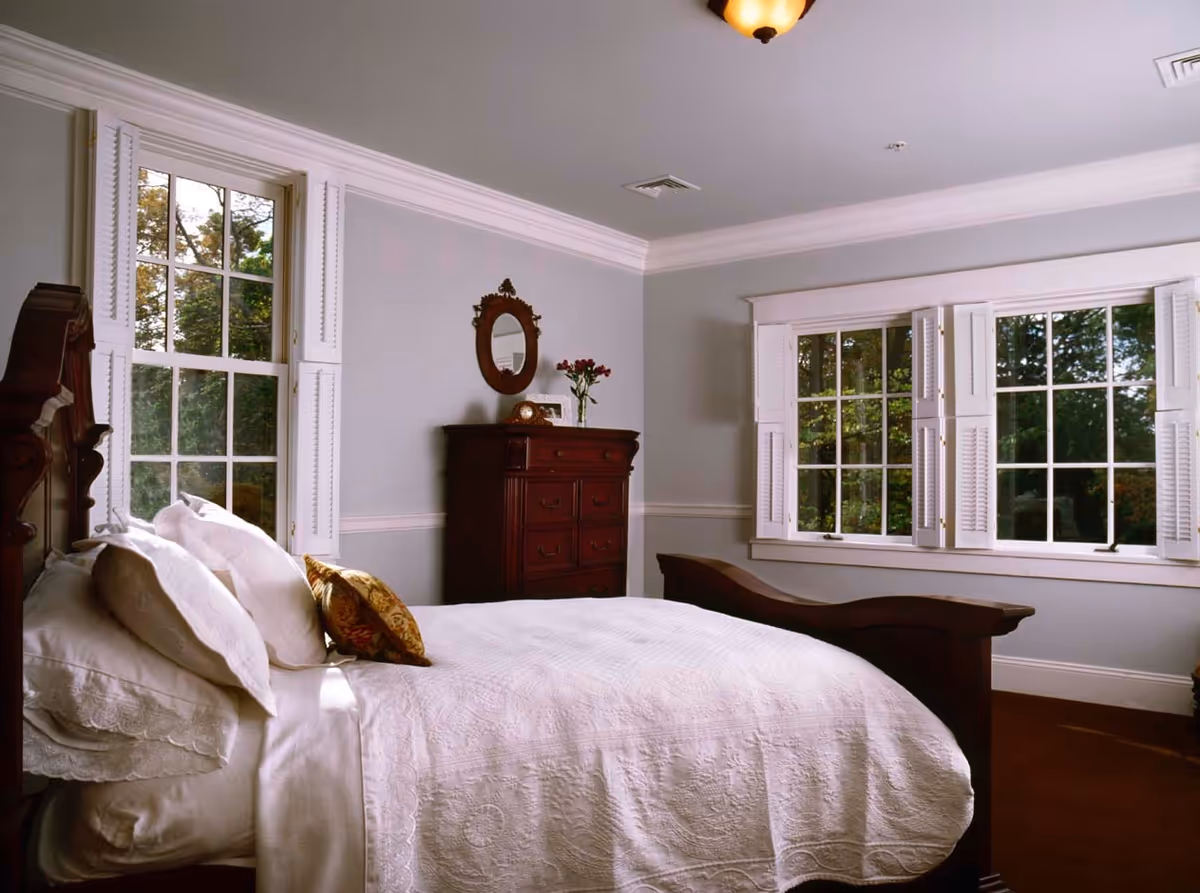 A cozy bedroom with a large wooden bed covered in white bedding and pillows. The room has light gray walls, two large windows with white shutters letting in natural light, and a dark wooden dresser with a small oval mirror and a vase of flowers on top.