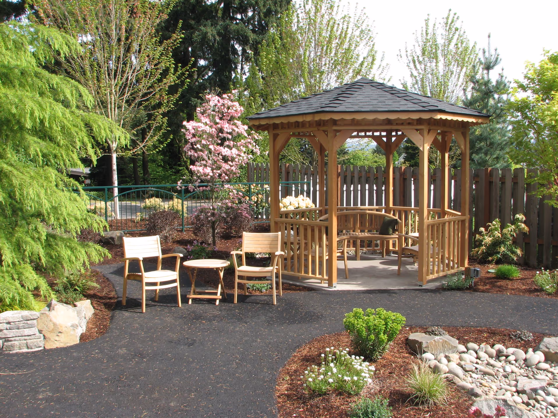 A peaceful outdoor garden area with a wooden gazebo featuring a bench and a cushion inside. In front of the gazebo, there are two wooden chairs and a small round table on a paved pathway. The garden is surrounded by various green trees, shrubs, flowering plants, and a rock arrangement.