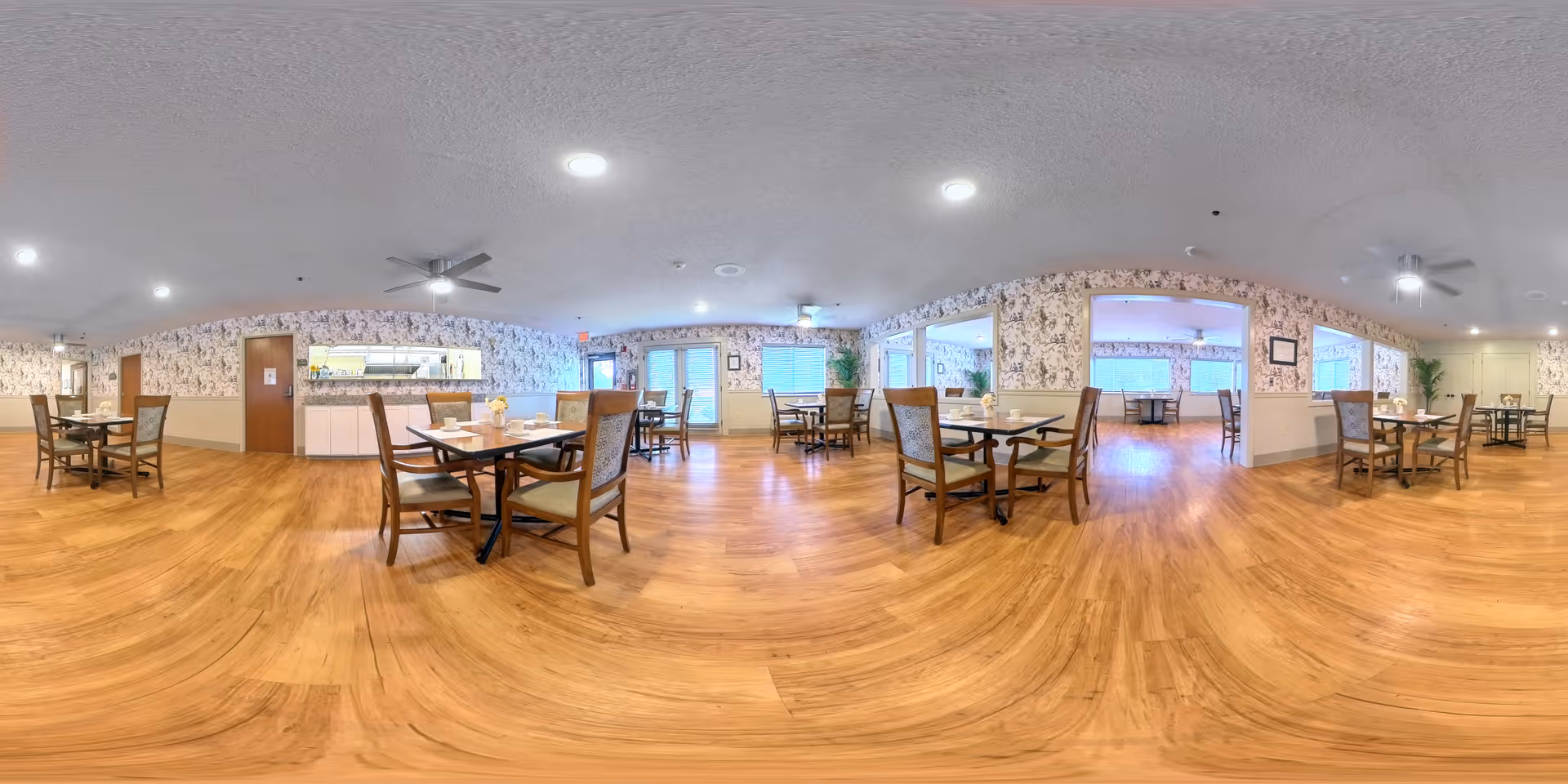 A spacious dining room with wooden floors and multiple tables and chairs arranged for seating. The walls have floral wallpaper and there are ceiling fans and recessed lights on the ceiling. Large windows allow natural light to enter the room.