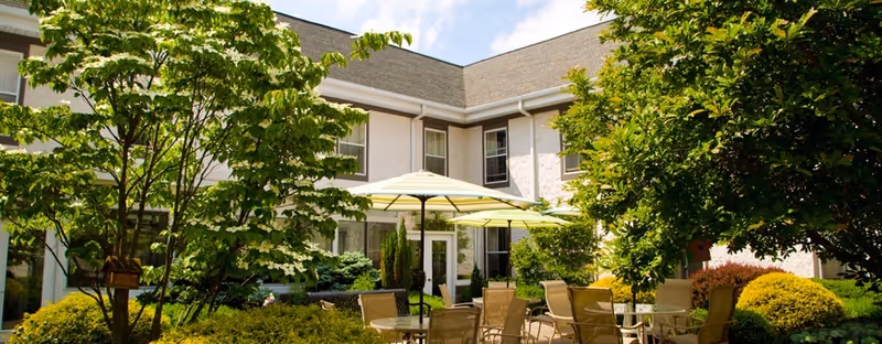 Sunny courtyard patio with tables, chairs and umbrellas surrounded by shrubs and trees in front of a two-story facility building.