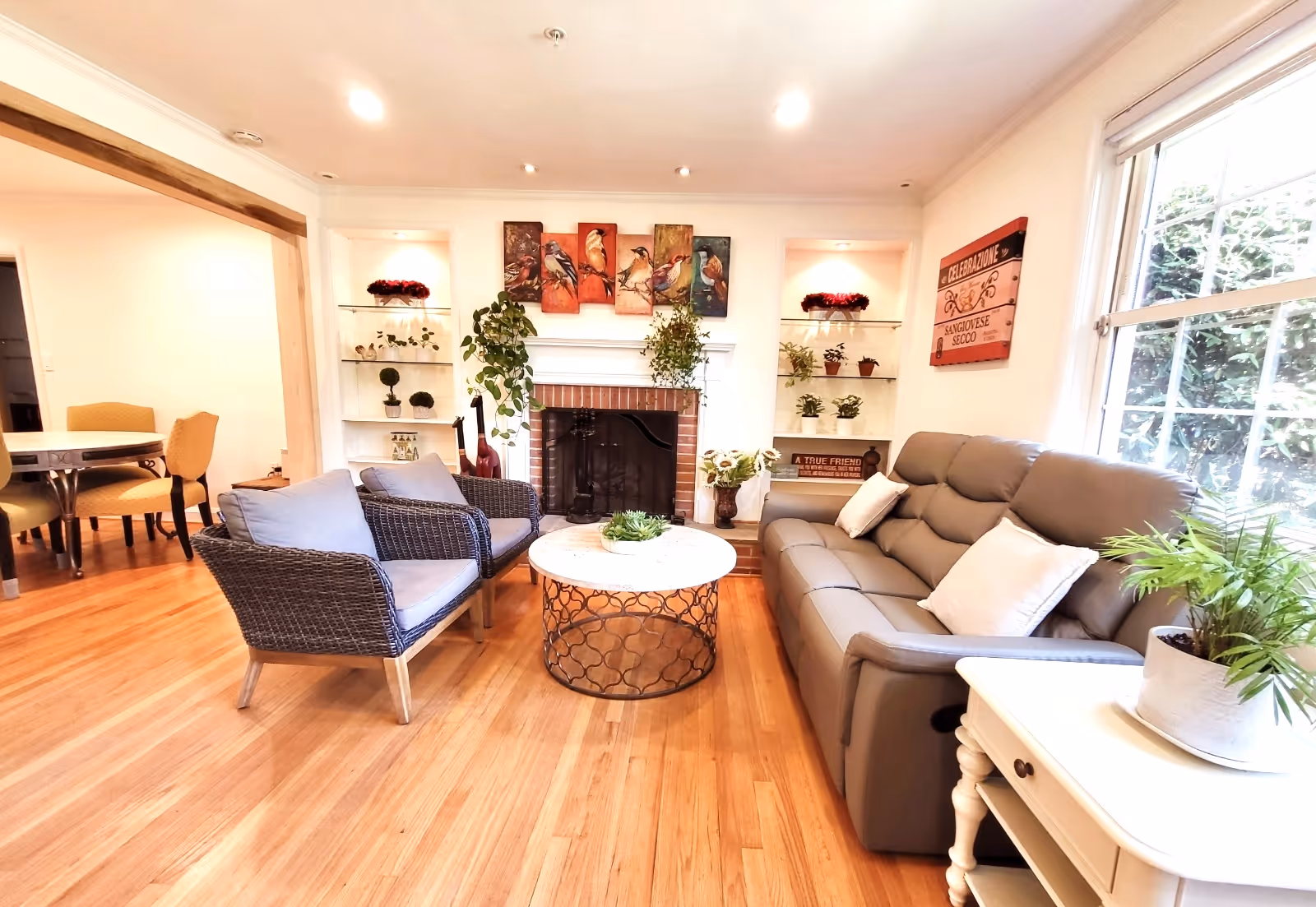A bright and cozy living room with wooden flooring, featuring a gray sofa with white pillows, two wicker armchairs with cushions, and a round coffee table with a decorative plant. The room has a fireplace with a bird-themed artwork above it and built-in shelves on either side holding various plants and decorations. A large window lets in natural light, and a small white side table with a potted plant is next to the sofa. In the background, a dining area with a round table and chairs is partially visible.