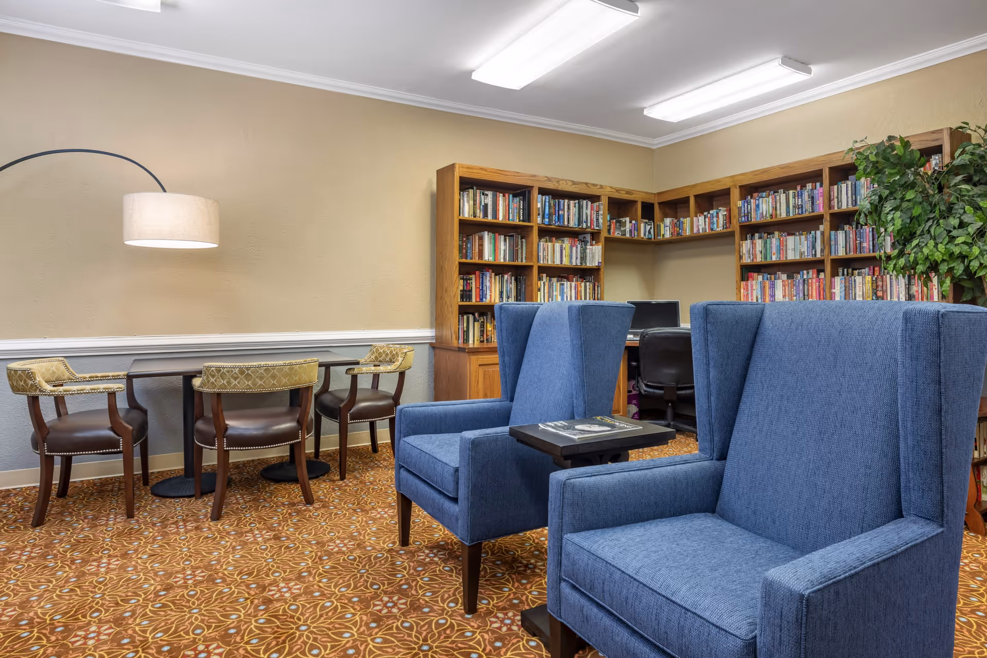 A cozy reading and seating area in a senior living facility featuring two blue upholstered armchairs with a small table between them, a corner bookshelf filled with books, a desk with a computer, and a table with four chairs under a large curved floor lamp. The room has patterned carpet and beige walls.
