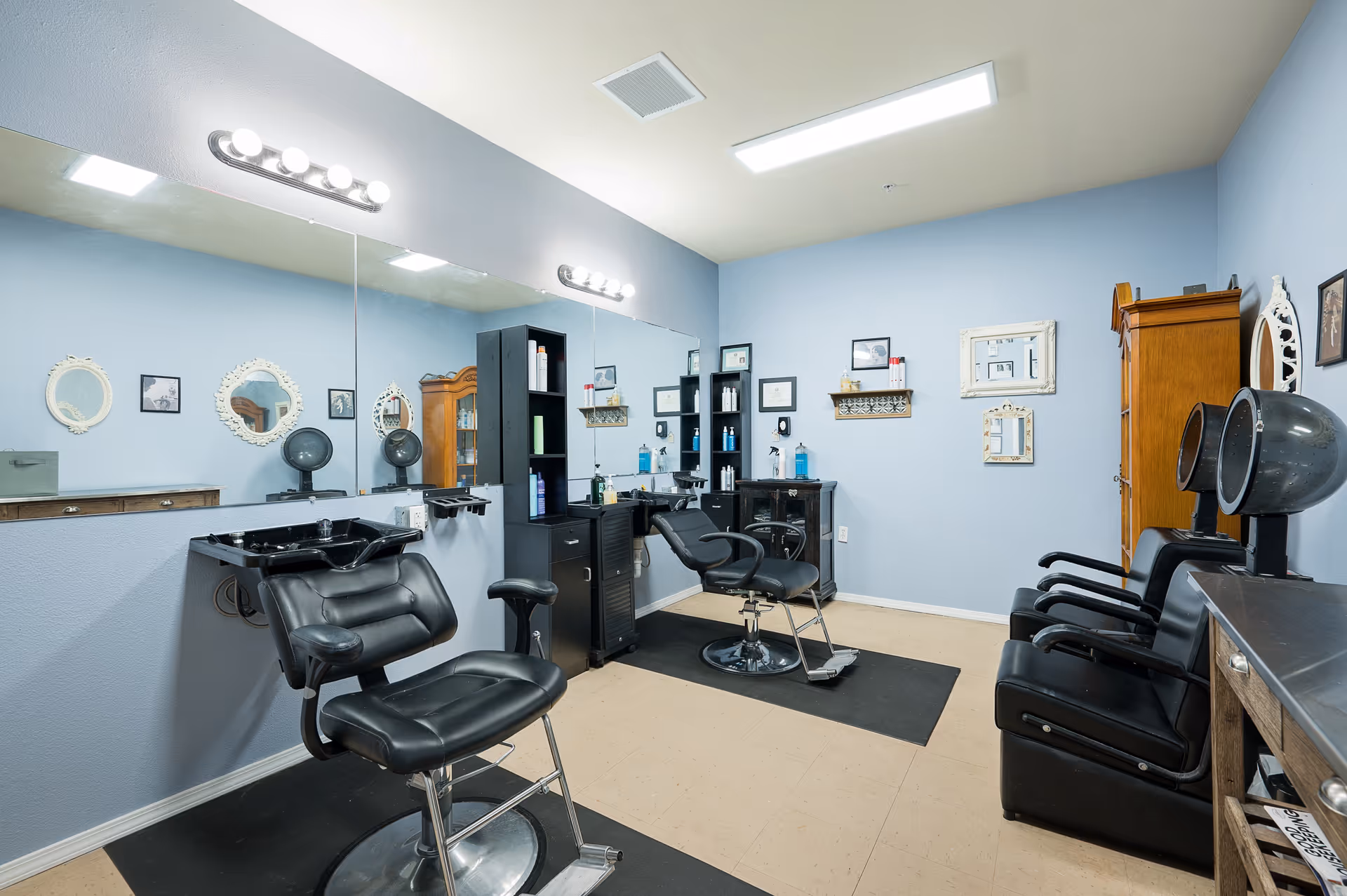 Interior of a salon room with two black salon chairs in front of a large mirror, hair washing sinks, hair dryers, shelves with hair products, and light blue walls decorated with framed pictures and mirrors.
