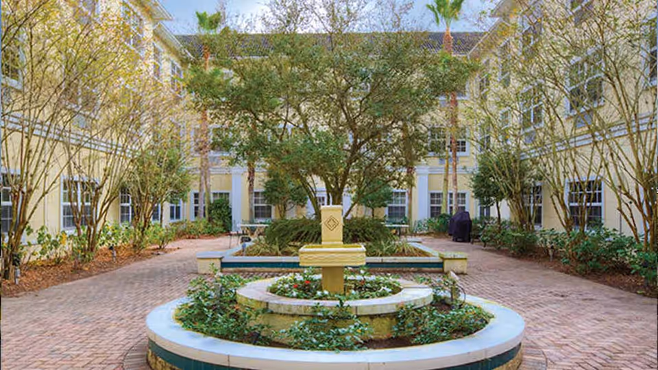 Sunny courtyard with a central circular fountain planter, paved walkways, trees and surrounding three-story yellow building.