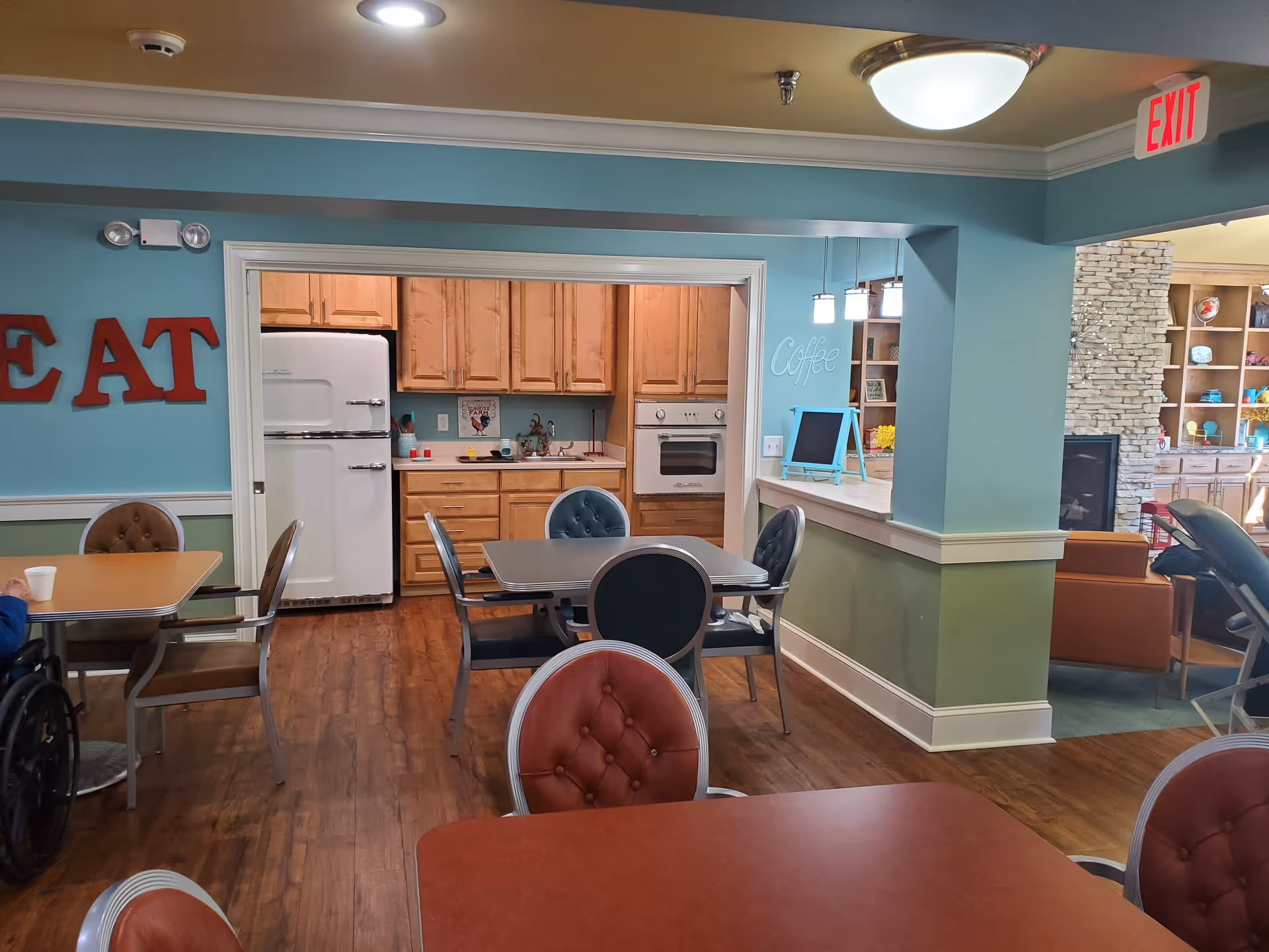 Interior view of a senior living facility showing a dining area with tables and chairs, a kitchen with wooden cabinets, a white refrigerator, and an oven. The walls are painted blue and green, with red letters spelling 'EAT' on the left wall and the word 'Coffee' on the right wall near a small chalkboard. In the background, there is a living room area with a stone fireplace and built-in shelves.