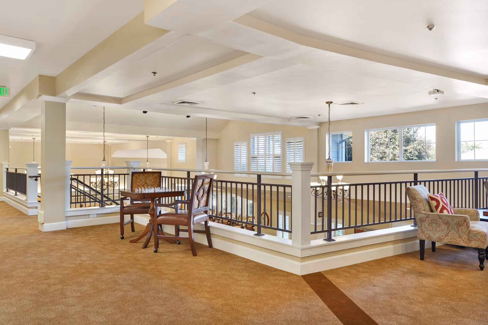 A bright and spacious indoor common area with beige walls and carpeted floor. There is a wooden table with two chairs, one of which has a chessboard on it. To the right, there is a patterned armchair with a red and white pillow. The area is bordered by a black metal railing overlooking a lower level with large windows letting in natural light.