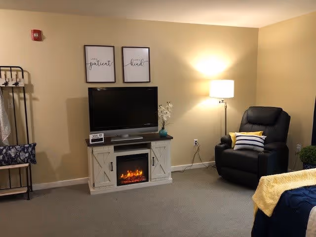 A cozy living room area with a black recliner chair featuring a striped pillow and a yellow pillow, a floor lamp emitting warm light, a TV on a white wooden stand with a built-in electric fireplace, and two framed wall art pieces above the TV that say 'lovely patient' and 'always kind'. A small plant and a decorative item are on the TV stand, and a bed with a yellow blanket is partially visible on the right side.
