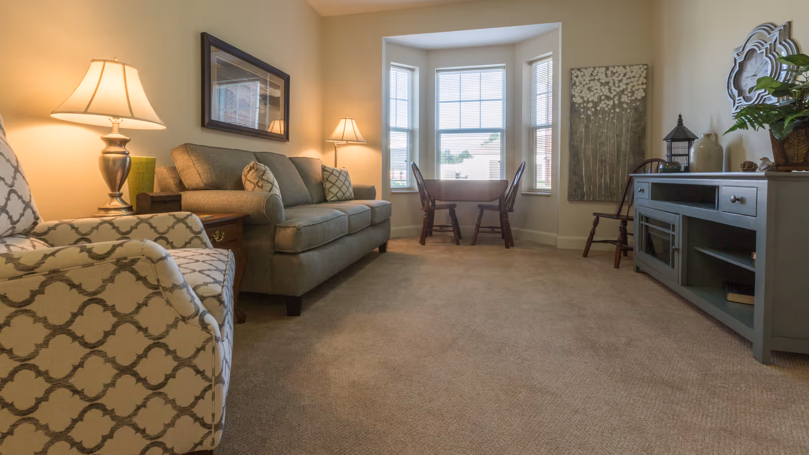 Comfortable living room featuring a patterned armchair and sofa, lamps, a media console, and a bay window with a small table and chairs.