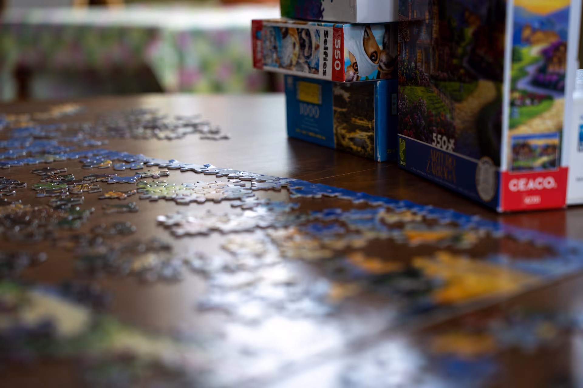 Partially assembled jigsaw puzzle pieces spread across a wooden table with stacked puzzle boxes at one end.