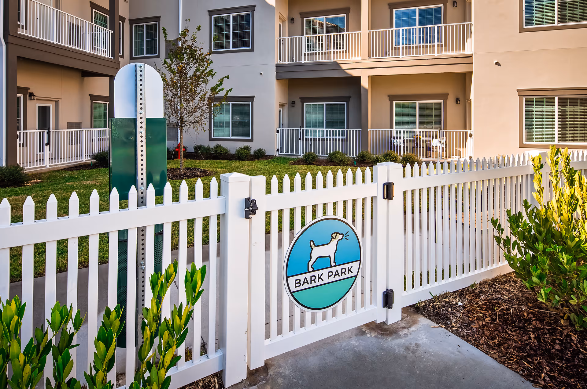 A fenced outdoor area labeled as a 'Bark Park' with a white picket fence and a sign featuring a barking dog illustration. The background shows a multi-story residential building with balconies and windows, surrounded by green grass and small shrubs.