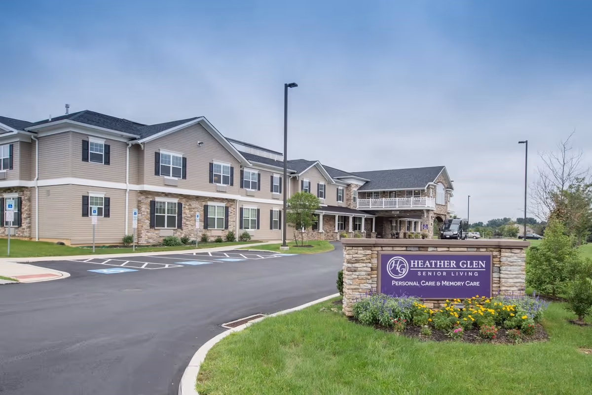 Exterior view of Heather Glen Senior Living facility showing a two-story building with beige siding and stone accents, surrounded by a paved driveway and landscaped greenery. A sign in front reads 'Heather Glen Senior Living Personal Care & Memory Care'.