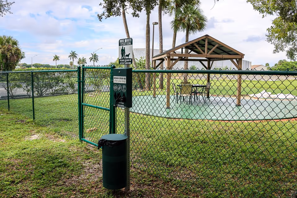 Outdoor fenced area with a dog waste station sign and trash bin attached to the fence. Inside the fenced area, there is a covered pavilion with a table and chairs. Palm trees and a grassy field are visible in the background under a partly cloudy sky.