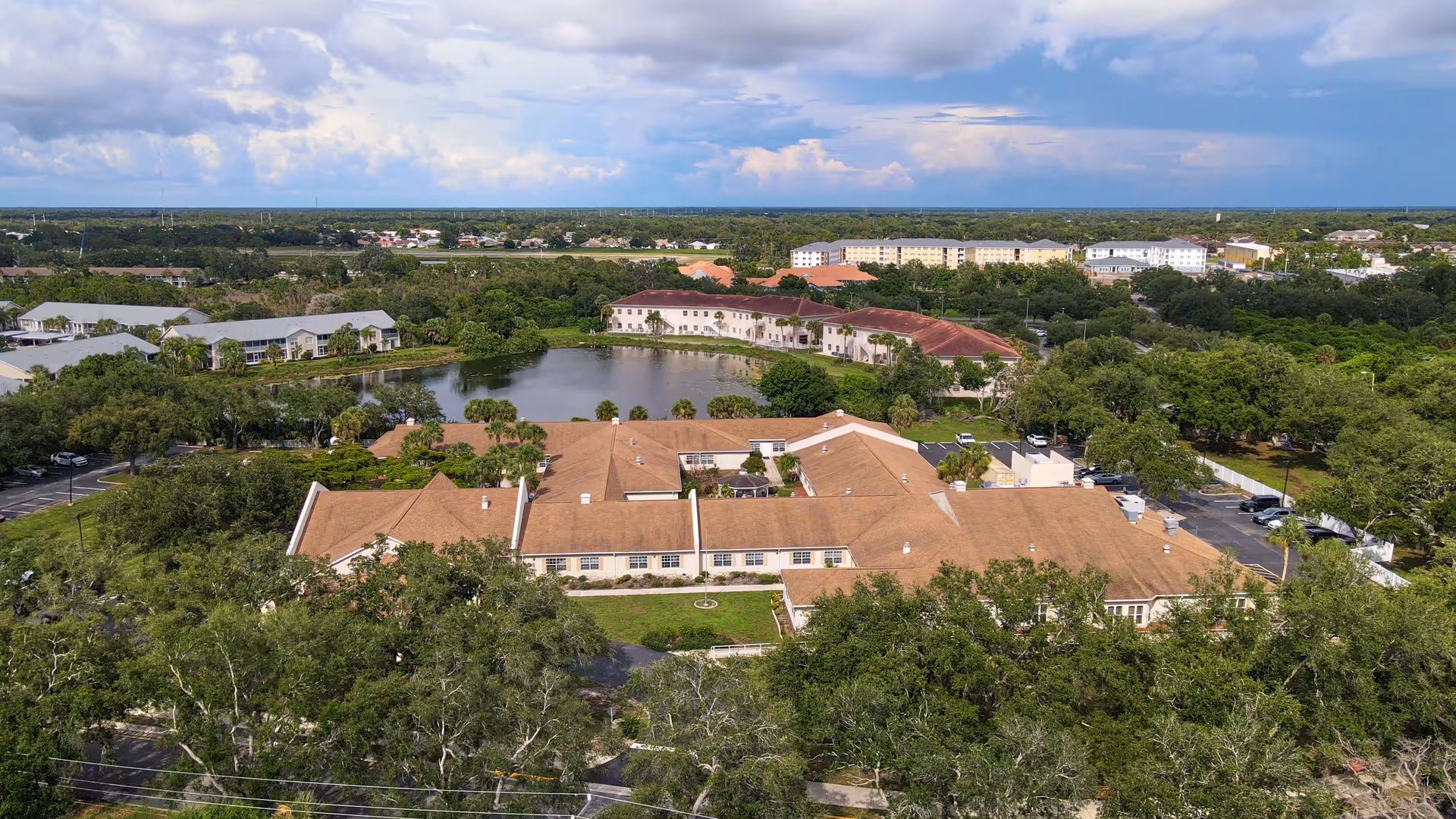 Aerial view of Sunset Lake Health and Rehabilitation Center surrounded by trees and greenery, with a pond and other buildings visible in the background under a partly cloudy sky.
