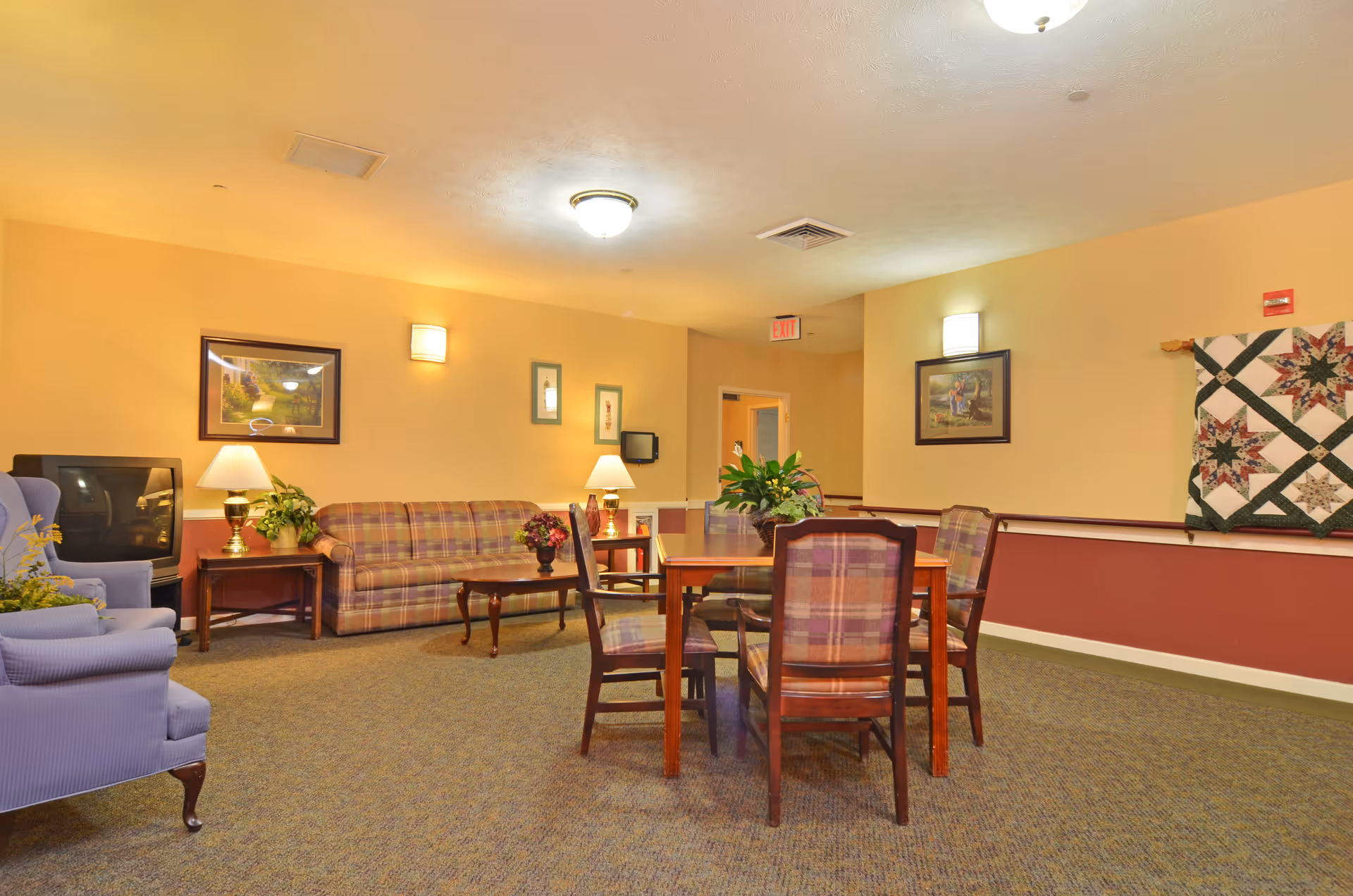 A cozy common area with a plaid sofa, a wooden coffee table, and a wooden dining table with four plaid chairs. There are two blue armchairs, a TV on a stand, framed pictures on the walls, table lamps, and a quilt hanging on the wall. The room has warm yellow walls and carpeted flooring.