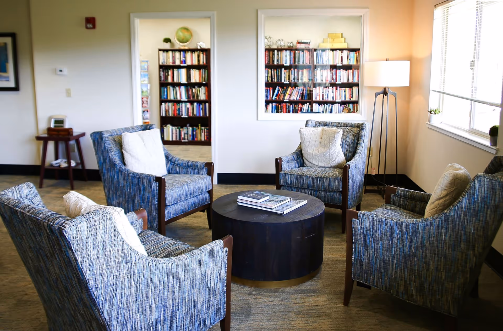 A bright lounge with four upholstered armchairs arranged around a round coffee table and a bookshelf visible through an interior window.