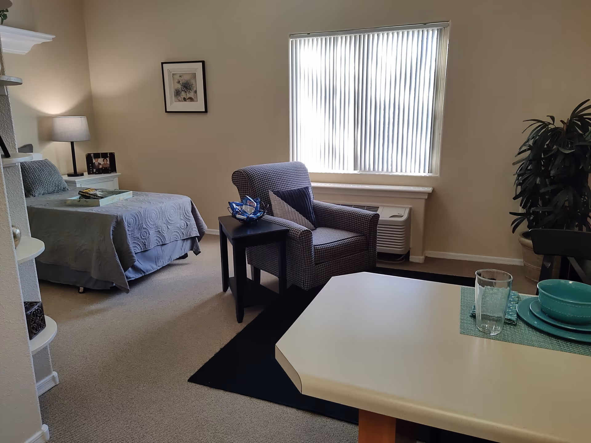 A cozy room with a single bed covered in a gray quilt, a nightstand with a lamp and a framed photo, a patterned armchair next to a small black side table, a large window with vertical blinds, a potted plant in the corner, and a dining table set with turquoise dishes and a glass.
