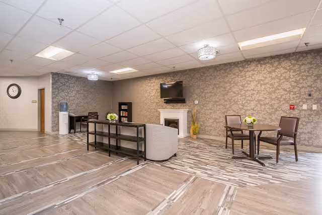 A spacious senior living common room with seating, small tables, a wall-mounted TV above a decorative fireplace, bookshelves, and patterned wallpaper.