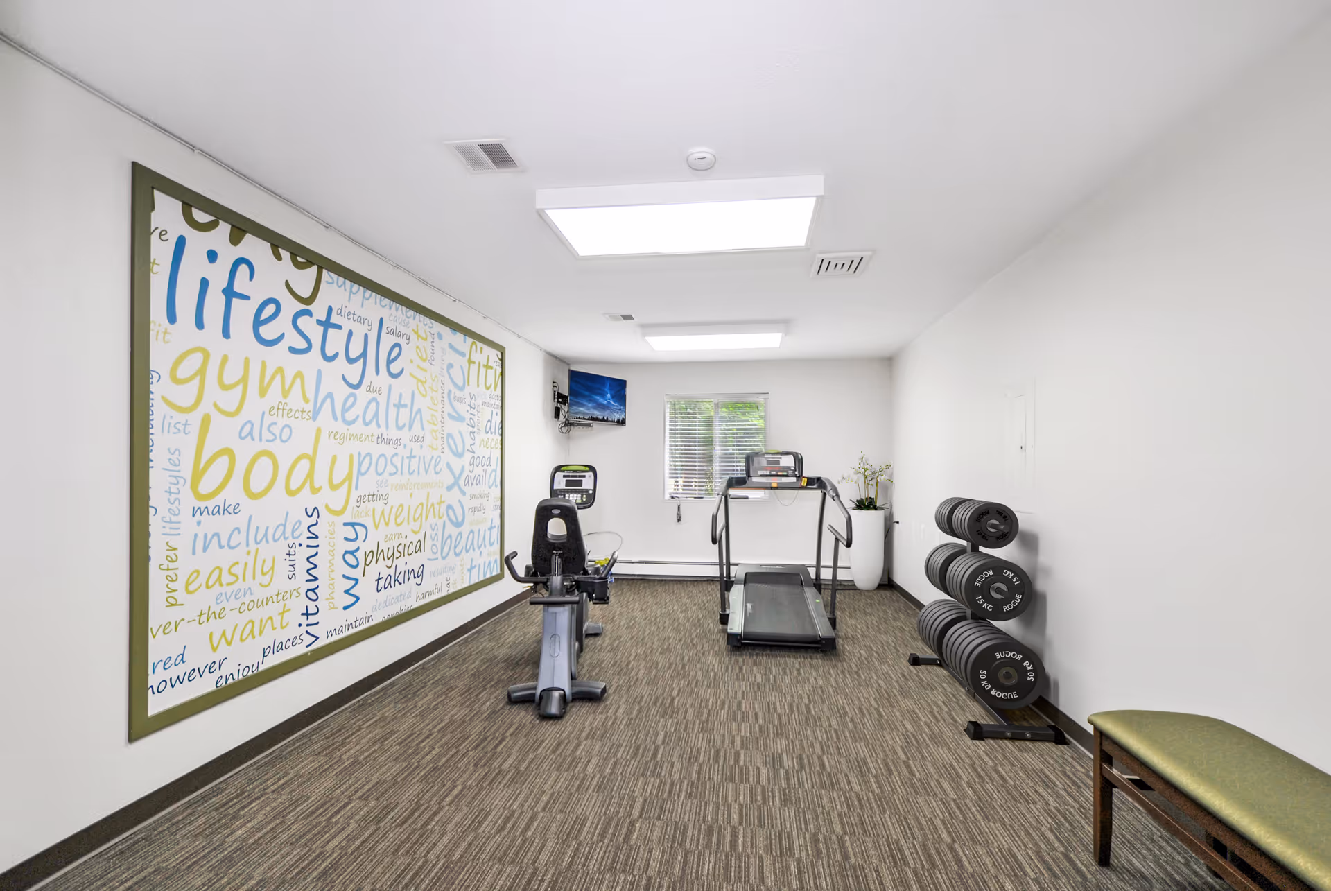 Small fitness room with a treadmill, recumbent bike, dumbbells, bench and a large word-art mural on the wall.