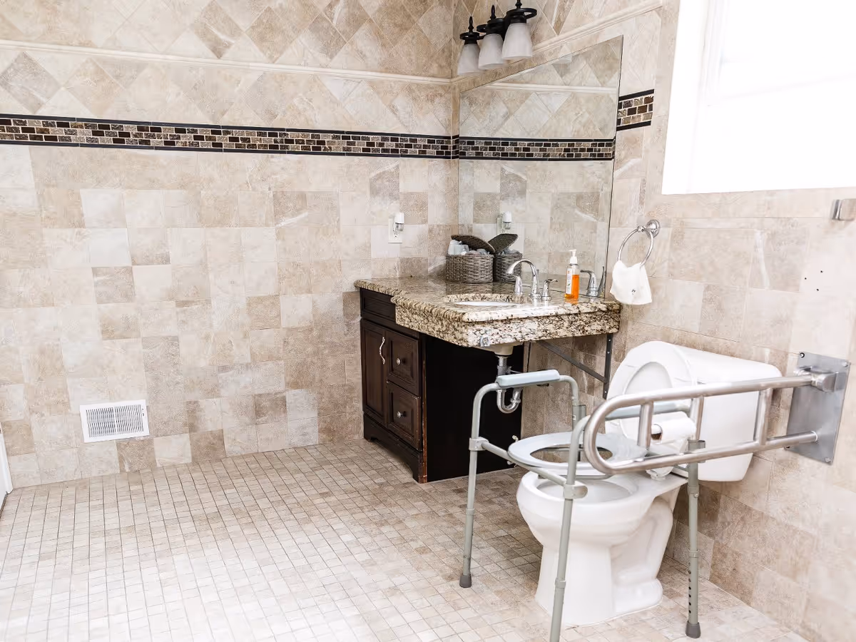 A spacious bathroom with beige tiled walls and floor. It features a granite countertop with a sink, a large mirror above the sink, and a white toilet equipped with safety rails and a walker frame for accessibility. There is a window allowing natural light into the room.