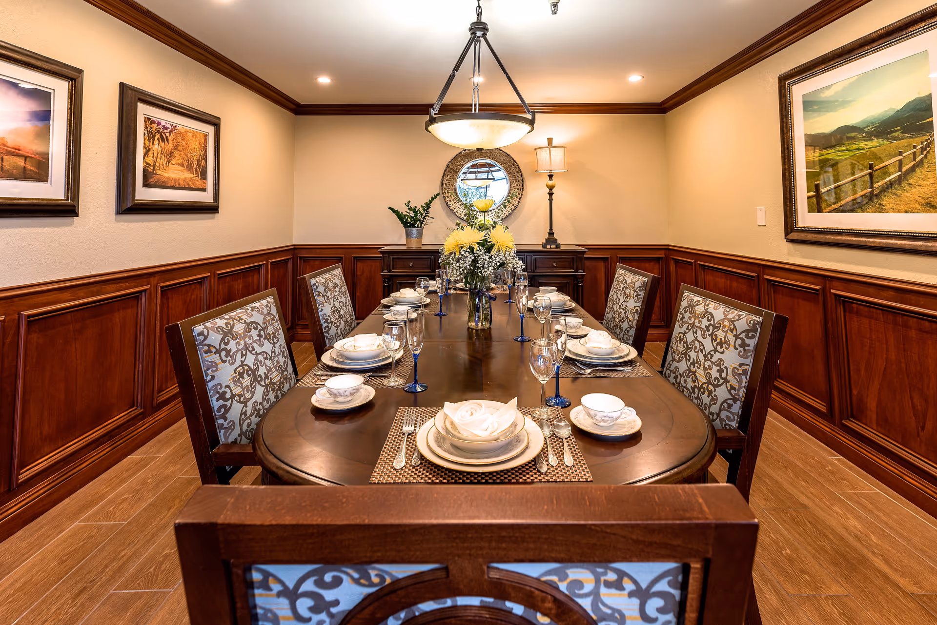 A formal dining room with a dark wooden table set for six people with white plates, bowls, and blue-stemmed glasses. The chairs have patterned upholstery. The room features wood paneling on the lower walls, beige upper walls, framed landscape paintings, a round mirror, a table lamp, and a hanging light fixture above the table.