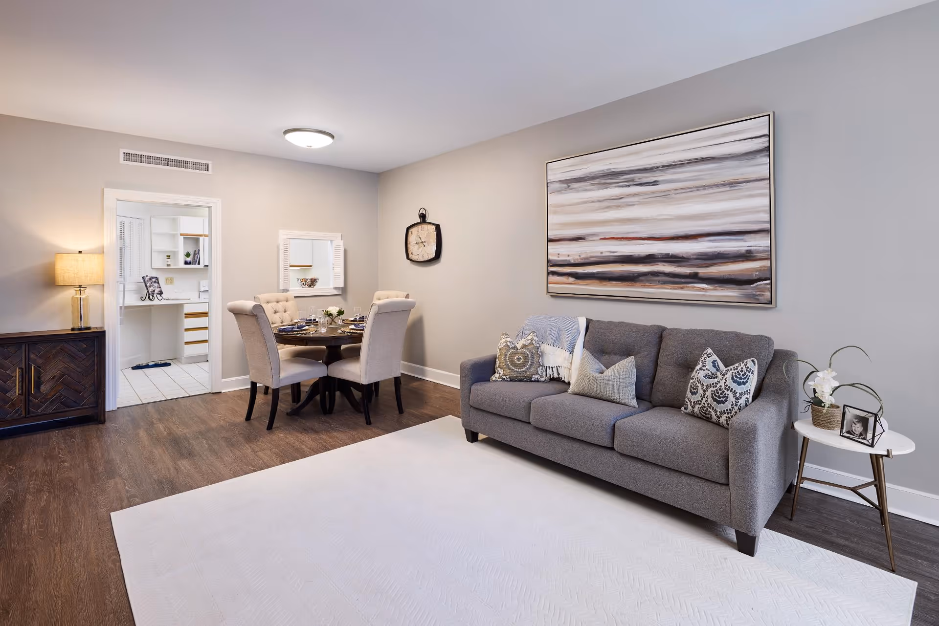 A modern living and dining area in a retirement community apartment. The room features a gray sofa with decorative pillows, a round dining table with four upholstered chairs, a wooden sideboard with a lamp, and a large abstract painting on the wall. The floor is dark wood with a large light-colored area rug. A doorway leads to a bright kitchen with white cabinets and tiled flooring.