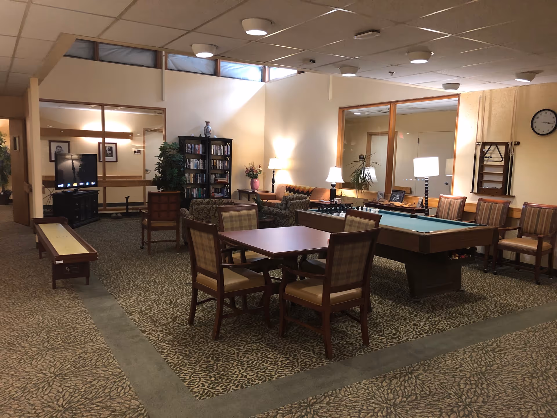 A cozy common area in a senior living facility featuring a pool table, a shuffleboard table, a chess set, a television, bookshelves filled with books, several chairs and tables, and soft lighting from lamps. The room has carpeted floors and large windows near the ceiling.
