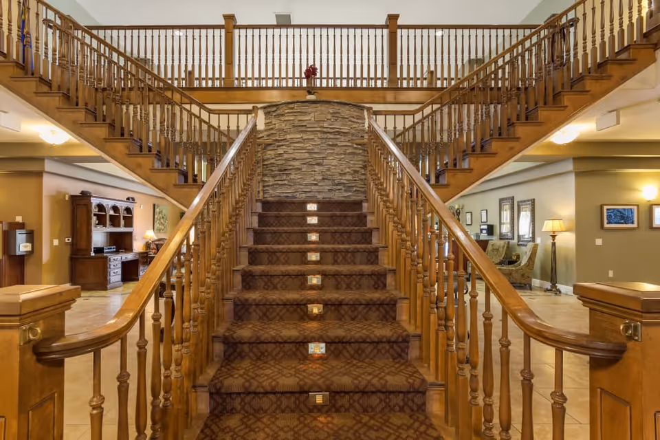 A wide wooden staircase with carpeted steps and wooden railings on both sides, leading up to a second-floor balcony with matching wooden balusters. The staircase is centered against a stone accent wall. On either side of the staircase, there are open interior spaces with furniture, lamps, and framed pictures on the walls, creating a warm and inviting atmosphere.