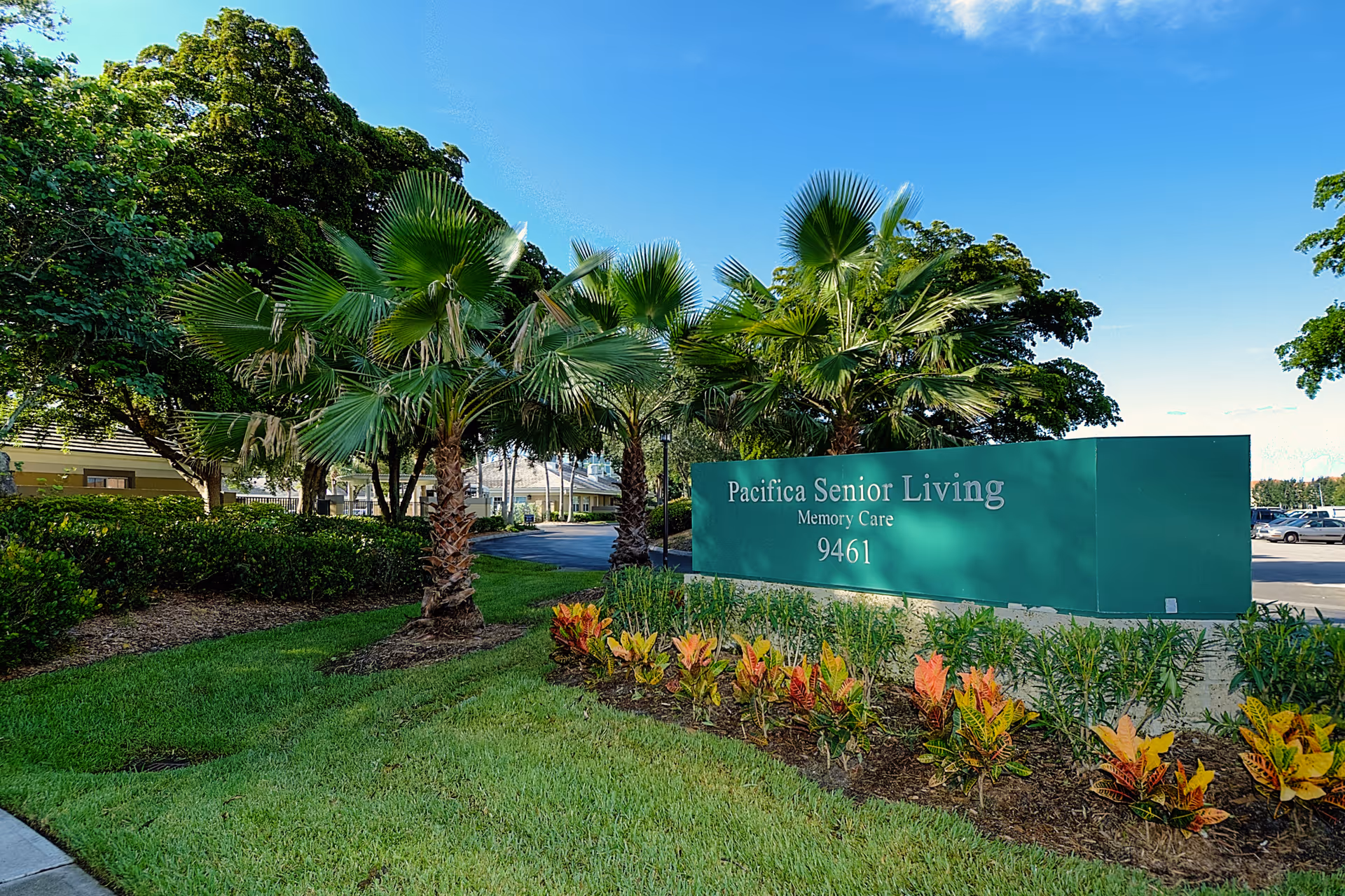 Outdoor view of the entrance area to Pacifica Senior Living Memory Care facility, featuring a green sign with the facility name and number 9461, surrounded by palm trees, bushes, and colorful plants under a clear blue sky.
