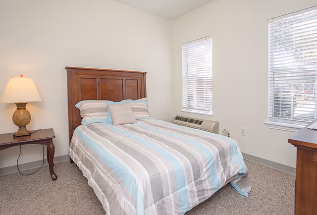 A neatly made bed with striped bedding in shades of blue, gray, and white in a simple bedroom. The room has two windows with white blinds, a wooden nightstand with a lamp, and a wooden dresser. The walls are white and the floor is carpeted.