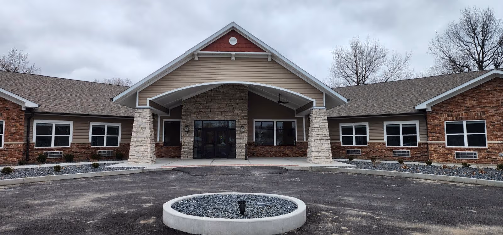 Front entrance of a single-story assisted living building with a covered portico supported by stone pillars and a circular planter in the driveway.