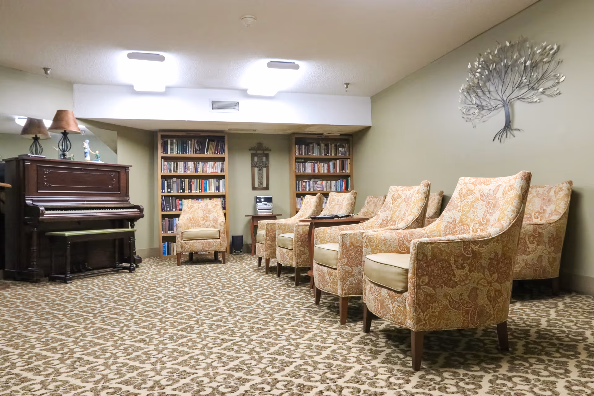 Carpeted lounge with patterned armchairs in a row, a piano to the left, and bookshelves along the back wall.