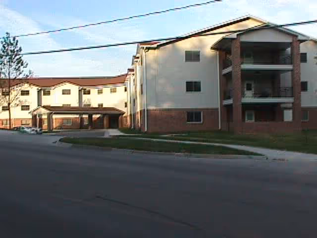 Exterior view of a multi-story senior living facility building with a covered entrance, balconies, and a paved road in front under a partly cloudy sky.