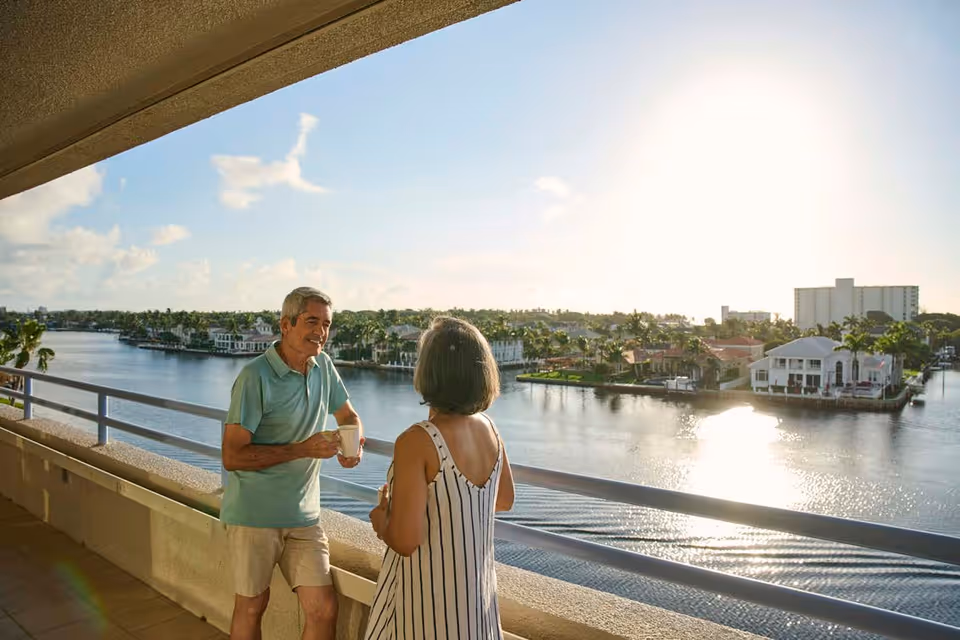 An older man and woman stand on a balcony overlooking a calm waterway with houses and palm trees on the opposite shore. The man is holding a coffee mug and smiling while talking to the woman, who is wearing a striped dress. The sun is shining brightly in the sky, creating a warm and peaceful atmosphere.