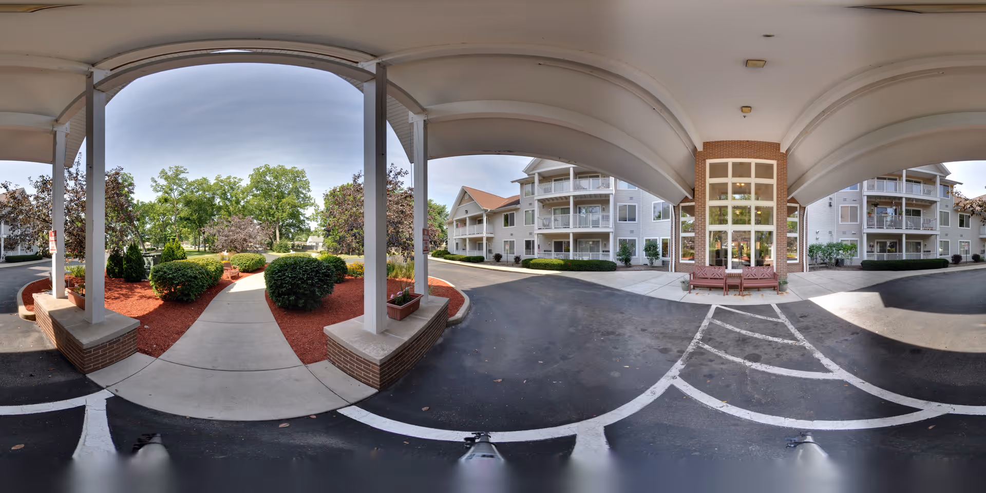 Covered entrance area of a senior living facility with a paved driveway and landscaped garden beds featuring bushes and trees. The building has multiple floors with balconies and large windows. Two benches are placed near the entrance under the covered area.